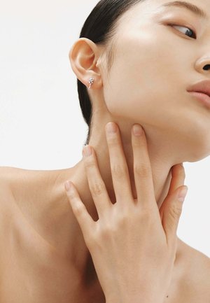 Pendiente de plata con una forma única y pequeños elementos decorativos; modelo con piel suave y un fondo neutro, sosteniendo suavemente el cuello.