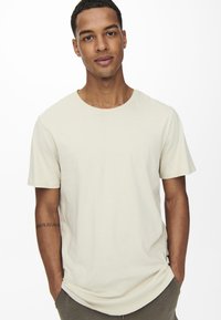 Cremfarbiges, kurzärmliges T-Shirt in lockerer Passform; aus weichem Stoff mit Rundhalsausschnitt; minimale Nähte; getragen mit den Händen in den Taschen.