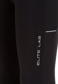 Gros plan sur un legging de sport noir avec l'inscription "ELITE LAB" en blanc sur la cuisse, équipé d'un panneau en mesh et d'une bande réfléchissante sur le côté.