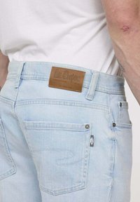 Jean en denim bleu clair avec une patch en cuir sur la taille, des poches arrière et des détails de coutures contrastés.