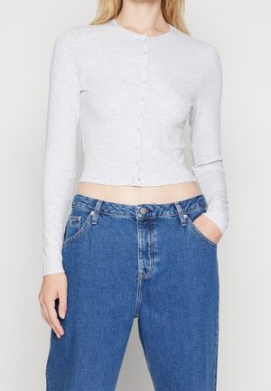 Kobieta w dopasowanym, długim rękawie, jasnoszarym krótkim koszulowym topie zapinanym na guziki i wysokim stanem niebieskich dżinsów, z jedną ręką w kieszeni.