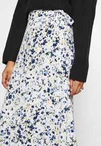 Jupe portefeuille florale blanche avec des motifs bleus, verts et jaunes. Dotée d'un détail à nouer à la taille. Tissu doux et design fluide.