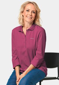 Magenta longsleeve polo met een puntige kraag, een drieknoopsluiting en subtiele logodetails. Gekleed met blauwe jeans.