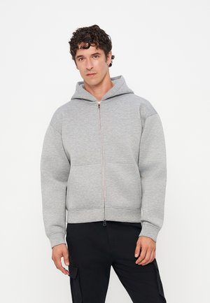 Young man with curly hair wearing a light gray zip-up hoodie and black cargo pants against a plain white background.
