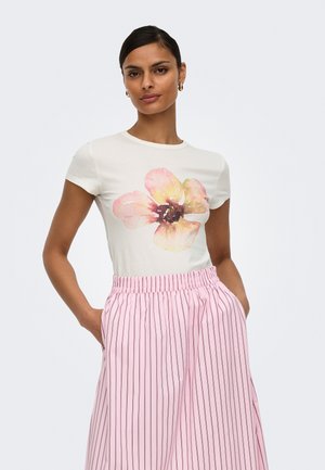 Nő fehér pólóban, rajta egy nagy rózsaszín és sárga virágmintás nyomat, rózsaszín-fehér csíkos bő szárú nadrággal párosítva, kezek a zsebekben.