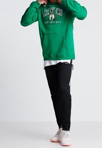 Zielona bluza z kapturem z grafiką "CELTICS BASKETBALL". Czarne spodnie z boczną lamówką. Jasne sneakersy dopełniają swobodny, sportowy wygląd.
