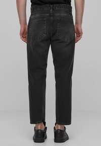 Zwarte denim jeans met een relaxte pasvorm, zichtbare stiknaden, twee achterzakken en een subtiele slijtage op de stof. Gemaakt met zwarte schoenen.