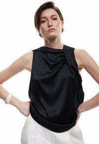 Mujer con cabello corto castaño, vistiendo una blusa negra de satén sin mangas y pantalones blancos, con las manos en las caderas, expresión neutral sobre un fondo blanco.