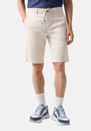 Mann trägt beige Shorts mit Kordelzug, weiße Kniesocken und blau-weiße Sportschuhe, steht mit den Händen in den Taschen.
