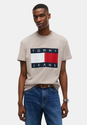 Joven con una camiseta beige de Tommy Jeans y vaqueros azules, con un cinturón marrón y un reloj plateado en la muñeca izquierda.