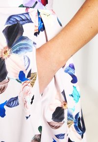 Blommigt klädestyg med livfulla blå, rosa och gröna mönster. Har en sidficka och en mjuk textur mot en ljus bakgrund.