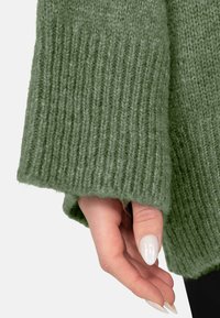 Mano con unghie lunghe bianche parzialmente coperte dalla manica di un maglione verde spesso a coste.