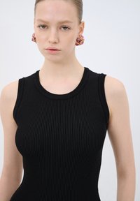 Černé žebrované bezramínkové šaty s kulatým výstřihem. Modelka má na sobě růžové výrazné náušnice se zlatými akcenty. Hladká textura, těsný střih.