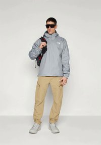 Light gray waterproof jacket with a zip, beige cargo pants, gray shoes, and a black and red backpack, accessorized with dark sunglasses.