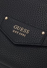 Juodas tekstūruotas dirbtinės odos maišelis su auksinės spalvos "GUESS EST 1981" logotipo plokštele ir mažomis metalinėmis detalėmis. Paprastas geometrinis dizainas.