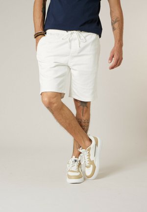 Man draagt witte shorts met trekkoord, beige en witte sneakers, een marineblauw shirt en kralenarmbanden, staat met gekruiste benen.
