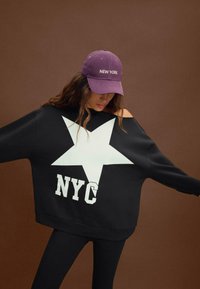 Felpa nera oversize con una grafica a forma di stella bianca e testo "NYC". Abbinata a un cappellino viola con la scritta "NEW YORK" in bianco.