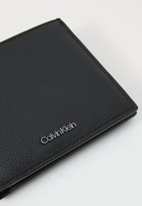 Schwarzes Lederportemonnaie mit strukturiertem Oberflächen, ausgestattet mit silbernem "Calvin Klein"-Logo. Kompakte rechteckige Form, saubere Kanten, dezente Nähte.