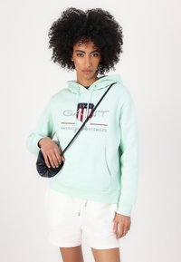 Mintgroene hoodie met "GANT" logo, trekkoord capuchon en kangoeroezak. Gemanierd met witte shorts en een kleine zwarte tas.