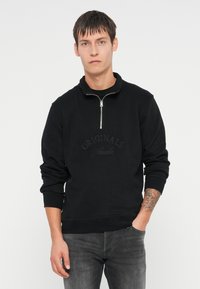 Sudadera negra con cuello de media cremallera, que presenta el texto en relieve "ORIGINALS Studio" en la parte frontal, mangas ajustadas y un corte relajado.