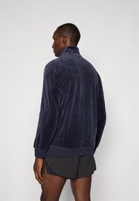 Tumedat sinine fliiskampsuni kõrge kraega, soonikkoes randmete ja äärega. Pehme tekstuur, sile viimistlus ja minimalistik disain.