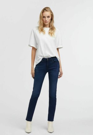 Donna bionda che indossa una t-shirt bianca larga, jeans skinny blu scuro e stivaletti beige, in piedi davanti a uno sfondo bianco uniforme.