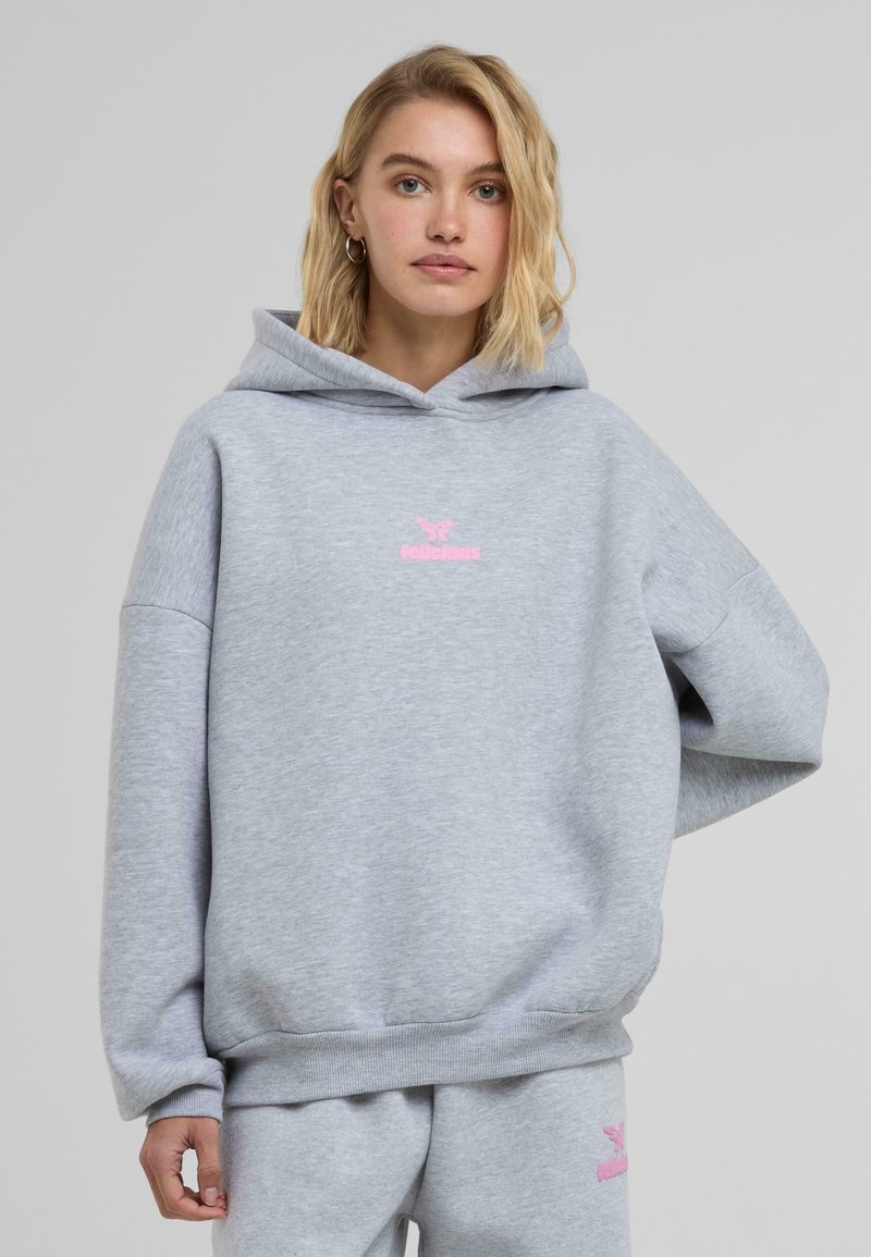 Valkoinen harmaa huppari, joka on valmistettu puuvillasekoitteesta ja jossa on vaaleanpunainen brodeerattu "felicious"-logo rinnassa. Huppari on löysähkössä istuvuudessa ja siinä on ribbaushelma.