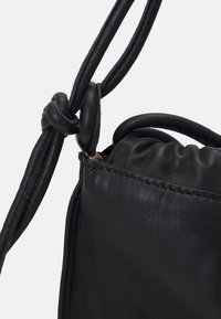 Gros plan d'un sac en cuir noir avec une bandoulière nouée et un bord supérieur froncé sur un fond clair uni.