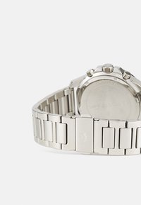 Reloj de acero inoxidable con una pulsera de plata pulida, que presenta eslabones rectangulares y una caja redonda. Logo grabado en el cierre.