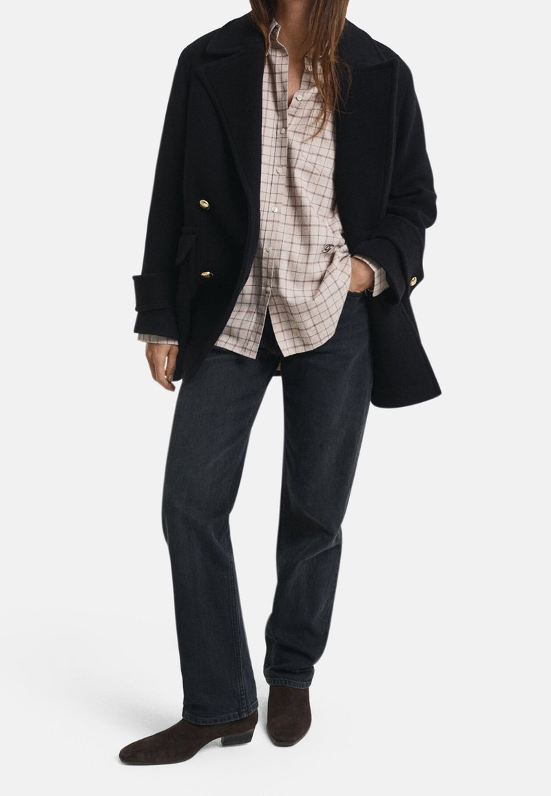 Personne portant un manteau noir avec des boutons dorés sur une chemise beige à carreaux, un jean foncé et des bottes à talons marron foncé, debout avec une main dans la poche.