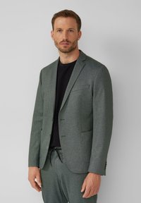 Veste de costume grise texturée avec une coupe ajustée, design à un seul boutonnage et revers crantés, associée à un T-shirt noir et un pantalon assorti.
