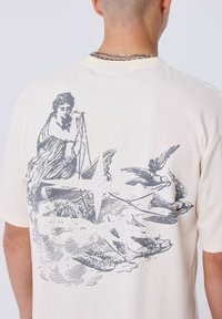 Camiseta color crema con estampado gráfico en la parte posterior, que presenta una figura sentada, aves y nubes en gris. Textura suave, corte holgado.