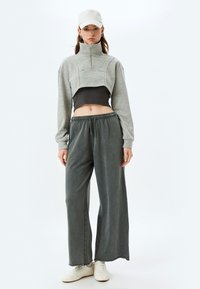 Grijze cropped zip sweatshirt over een donkere top, gecombineerd met losse, grijze wijdere broek en witte sneakers, met een baseballcap.