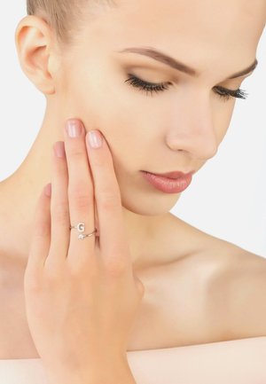 Silver ring featuring a "G" accent and a small stone. Model gently touches her cheek with her hand, showcasing the ring clearly.