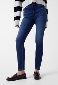 Jean slim en denim bleu foncé avec une taille haute, présentant des lignes épurées et un minimum de détails métalliques. Associé à des mocassins foncés et élégants.