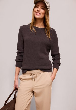 Kvinde iført en mørkebrun sweater, beige bukser med snøre, leopardmønstret kasket og med en brun taske mod en ensfarvet baggrund.