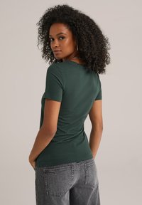 Vrouw met krullend haar in een donkergroene, aansluitende t-shirt en vervaagde zwarte jeans, staand met handen in haar zakken, kijkend over haar schouder.