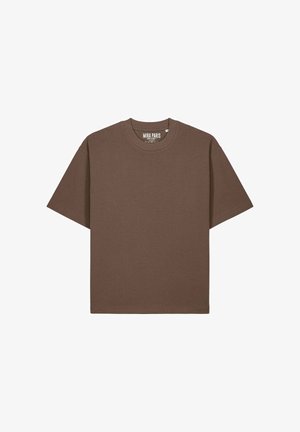 Donna con capelli neri fino alle spalle che indossa una maglietta marrone larga infilata in jeans blu a vita alta, in piedi contro uno sfondo semplice.