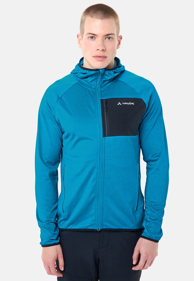 Jeune homme portant un sweat à capuche bleu avec une poche poitrine noire et le logo Vaude, debout devant un fond clair uni.