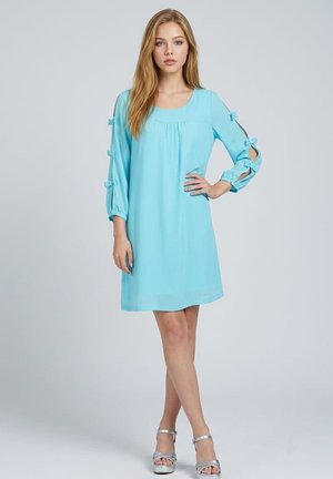 Joven mujer vestida con un vestido azul claro con mangas recortadas atadas con lazos, calzando sandalias plateadas de tacón alto sobre un fondo liso.