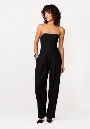 Kobieta o kręconych włosach, ubrana w czarny kombinezon bez ramiączek i srebrną bransoletę na rękę, stojąca na tle jednolitego białego tła.