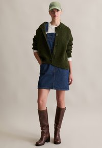 Cardigan en tricot vert olive, haut à manches longues blanc, robe salopette en jean bleu, et bottes en cuir marron jusqu'au genou ; look décontracté.