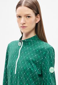 Πράσινο πουκάμισο με φερμουάρ και μοτίβο με κουκκίδες σε σχήμα καρδιάς σε διάφορες αποχρώσεις του πράσινου και του λευκού. Διαθέτει ψηλό γιακά και ετικέτα λογότυπου στο μανίκι.