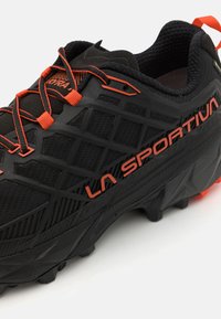Schwarzer Laufschuh mit strukturierter Oberfläche, orangefarbenen Akzenten und einer robusten Sohle. Besitzt ein Logo und ein filigranes Design im Obermaterial.