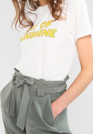 Kvinde iført hvid T-shirt med gul "Day of Sunshine" tekst og højtaljede olivengrønne bukser med et bundet stofbælte og hånd i lommen.