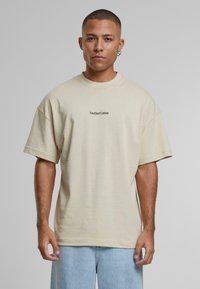 Kortærmet beigefarvet bomulds-t-shirt med en afslappet pasform og et lille sort "AnotherCotton" logo på brystet. Afslappet design og tekstur.