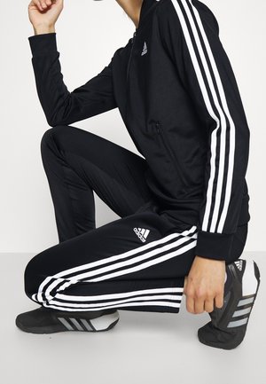 Osoba klęcząca, ubrana w czarny dres Adidas z białymi paskami i czarne buty sportowe Adidas, na jasnym, jednolitym tle.