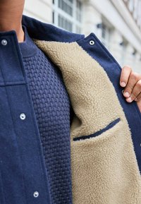Navy outer jacket with a soft beige fleece lining, featuring a textured knit sweater underneath and a small pocket in the lining.