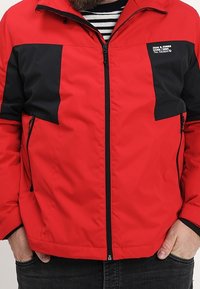 Rød lynlåstrøje med sorte accenter, med opretstående krave, to sidelommer og et mærket patch på brystet. Glat stoftekstur.