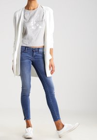 Cardigan blanco abierto por delante sobre una camiseta gris recortada. Jeans ajustados azules con cremalleras en los tobillos. Zapatillas blancas sin cordones. Atuendo simple y casual.